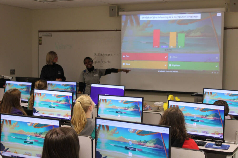 A classroom of students at desktop computers watches as two instructors lead an interactive quiz about computer languages, displayed on a projector at the front of the room.